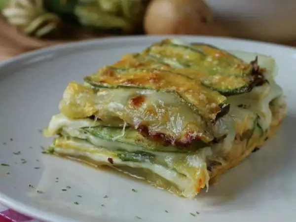 Una fetta di parmigiana di zucchine bianca, stratificata con formaggio filante e zucchine grigliate, servita su un piatto bianco.