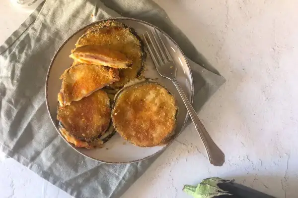 Cordon bleu di melanzane impanati e fritti, ripieni di formaggio filante e prosciutto, serviti su un piatto con una forchetta.