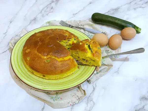Torta salata zucchine e speck dorata su piatto verde, con ingredienti freschi a lato.
