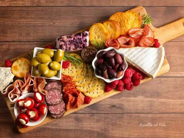 Un invitante tagliere salumi e formaggi con olive, lamponi, pane tostato e peperoncini ripieni.