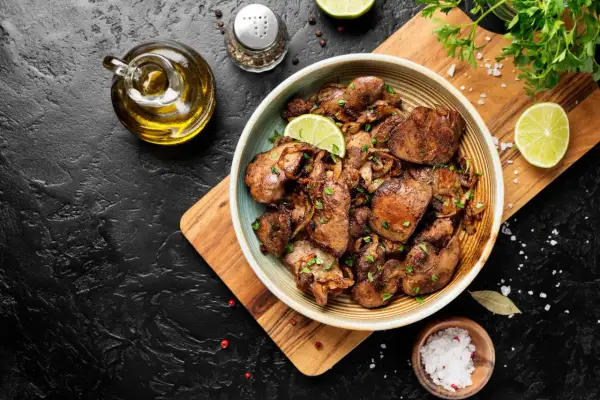 Piatto di fegatini di pollo saltati con cipolle, guarniti con prezzemolo fresco e spicchi di lime. Accanto, olio d'oliva, sale grosso e pepe.