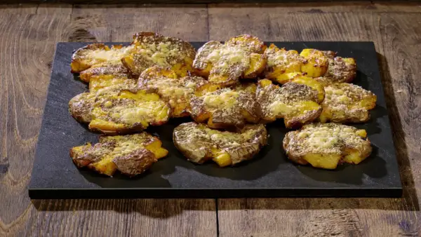 Patate schiacciate al forno, dorate e croccanti, cosparse di formaggio grattugiato e erbe aromatiche, servite su una pietra scura.